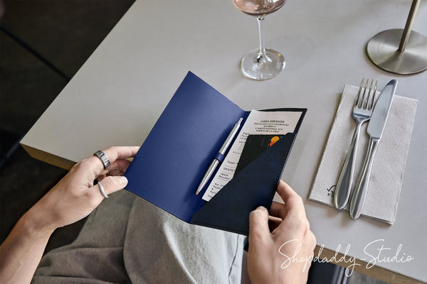A person holds an open navy blue folder or organizer with a pen and printed insert, seated at a light gray table with a wine glass, fork, knife, and napkin visible.