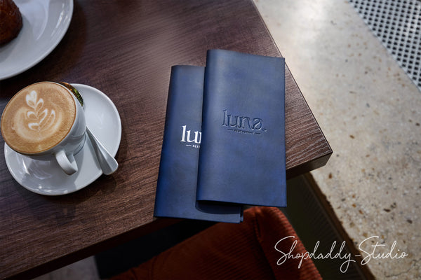 Two navy blue hardcover books or notebooks with embossed 'luna.' logos sit on a dark wooden table next to a cup of latte with latte art.