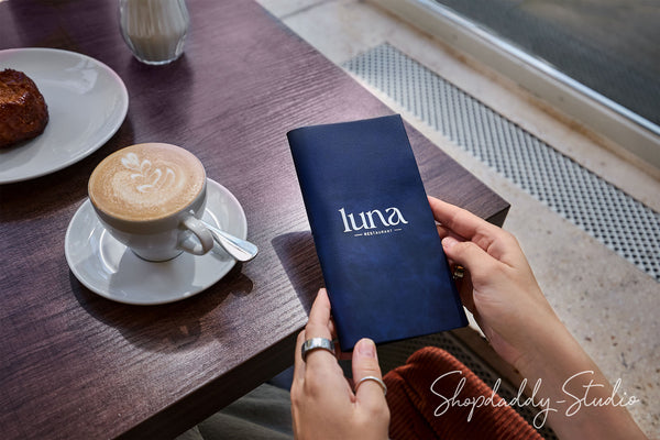 A person holding a navy blue menu or booklet with the word 'luna' printed in white, seated at a wooden table with a cup of latte and a pastry nearby.