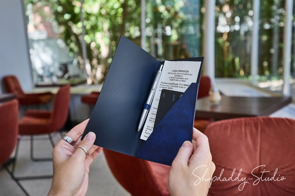 A person holds an open navy blue folder or organizer in a bright indoor setting with blurred red chairs and large windows in the background. A white card and pen are visible inside the folder.