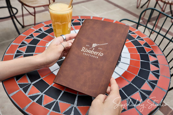 A person holds a brown paper menu for Rimberio Trattoria at an outdoor cafe table with a colorful mosaic pattern, next to a glass of orange drink.