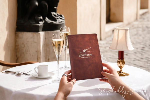 A person holds a brown leather-bound restaurant menu labeled 'Rimberto Trattoria' at an outdoor table setting with two champagne flutes, a white coffee cup, a table lamp, and warm sunlight.