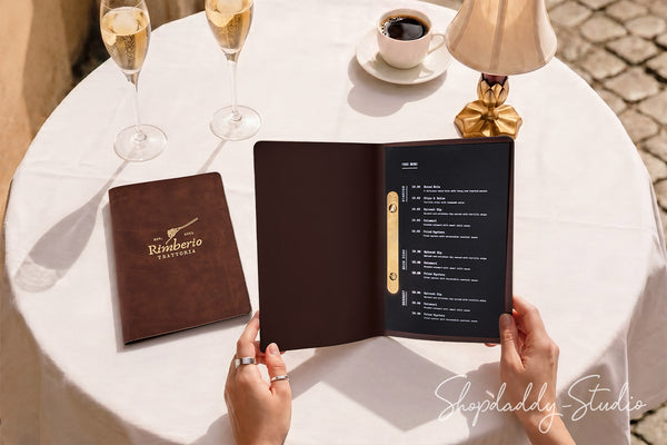 A person holds an open dark brown leather-bound restaurant menu with a gold ring binder, set on a white tablecloth beside a closed matching menu, two champagne flutes, a coffee cup, and a lamp.