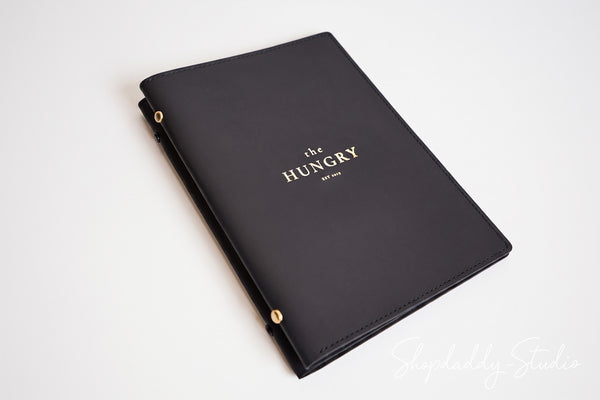 A black faux-leather menu cover with gold hardware and gold foil lettering that reads 'the HUNGRY' on a clean white background.