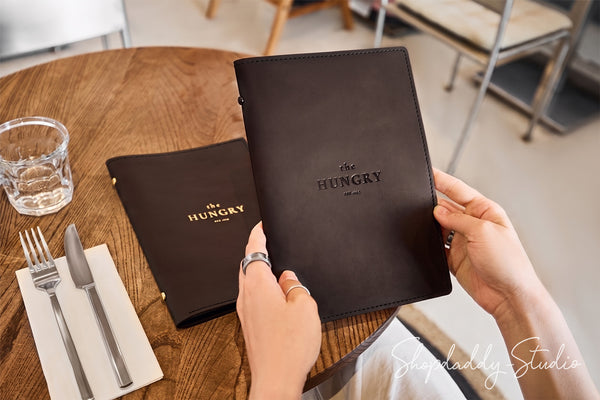 A person holds a dark brown leather menu from the restaurant 'The Hungry' at a wooden table, with another copy resting on the table next to a glass, cutlery, and napkin.