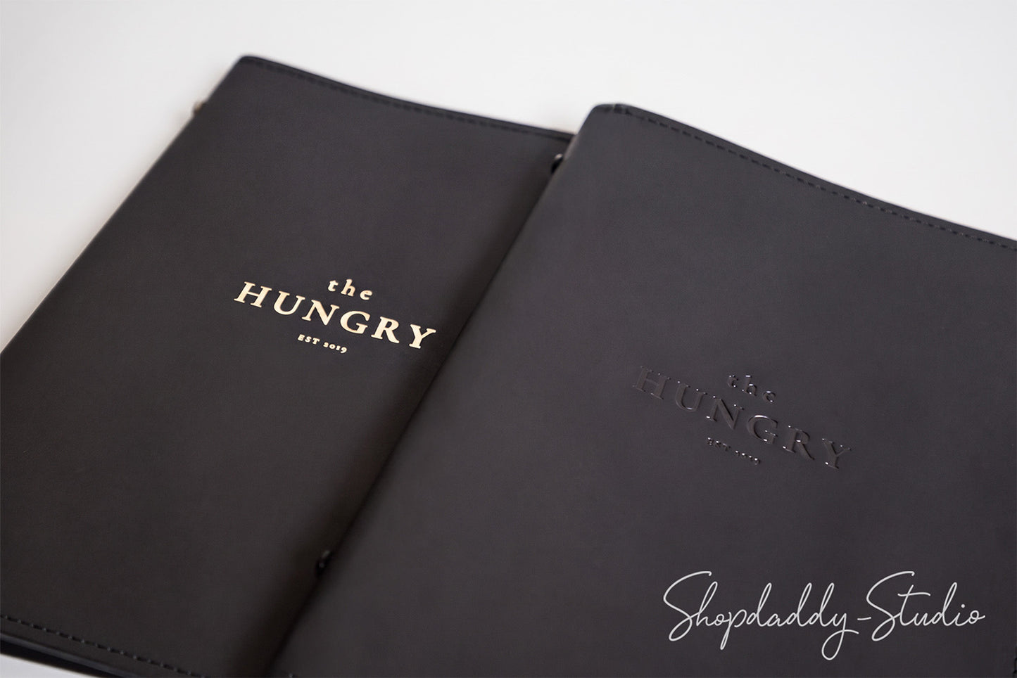 Two black leather planner covers with gold foil branding 'the HUNGRY EST. 2013' on a clean white background, with a Shopdaddy-Studio watermark in the corner.