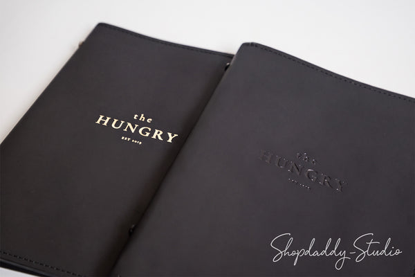 Two black leather planner covers with gold foil branding 'the HUNGRY EST. 2013' on a clean white background, with a Shopdaddy-Studio watermark in the corner.