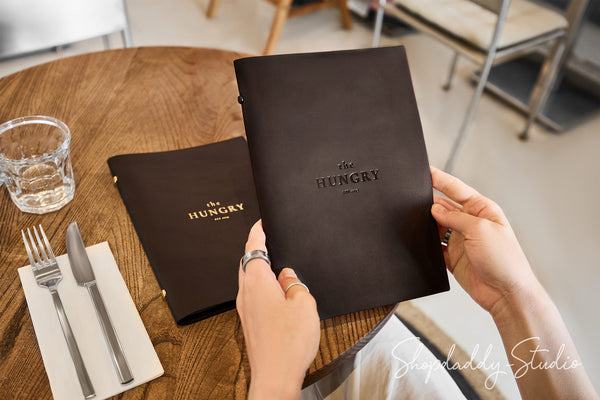 A person holds a dark brown leather menu from 'The Hungry Cafe' at a wooden table, with another copy of the menu, a glass, and silverware nearby.