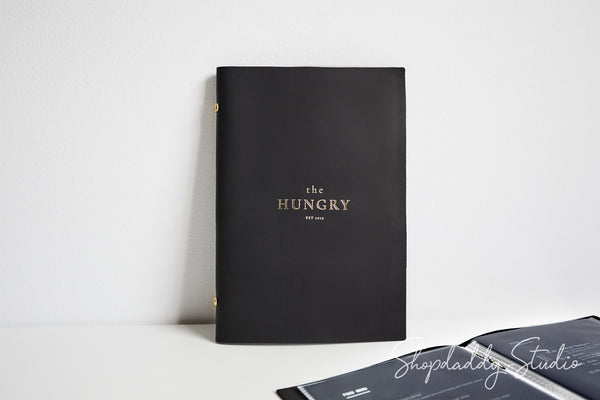 A black hardcover book titled 'the HUNGRY' with gold lettering, standing upright on a white surface against a white wall. A second black book or folder is partially visible in the lower right corner, and a Shopdaddy Studio watermark appears across the bottom.