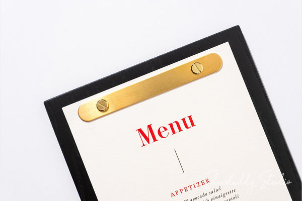 A close-up of a black-framed menu board with a gold metal clip and a white paper insert printed with the word 'Menu' in red, photographed on a light gray background.
