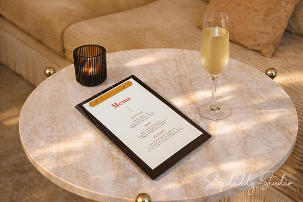 A black-framed menu with a gold accent band and white page sits on a round light marble coffee table beside a lit black candle and a glass of champagne, with a beige sofa in the background.