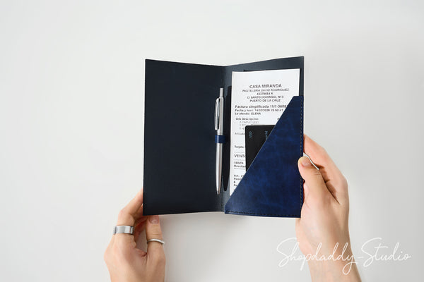 A person's hands holding open a navy blue passport cover or organizer, showing a white insert page, a silver pen, and a black phone inside. The photo is taken against a clean white background with a Shopdaddy Studio watermark in the corner.