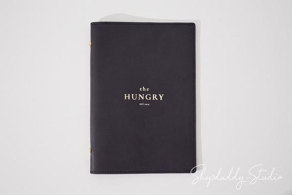 A black leather-bound journal with gold foil lettering that reads 'the HUNGRY EST. 2024' on the front cover, photographed on a clean white background.