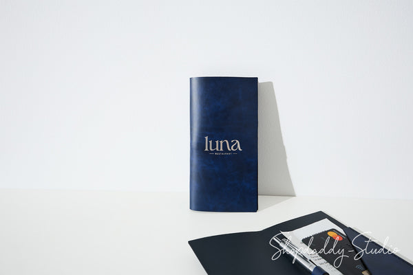 A dark blue textured notebook with the brand name 'luna' printed in white on the front, standing upright on a white surface against a white background. In the foreground, a black notebook and pen are partially visible.