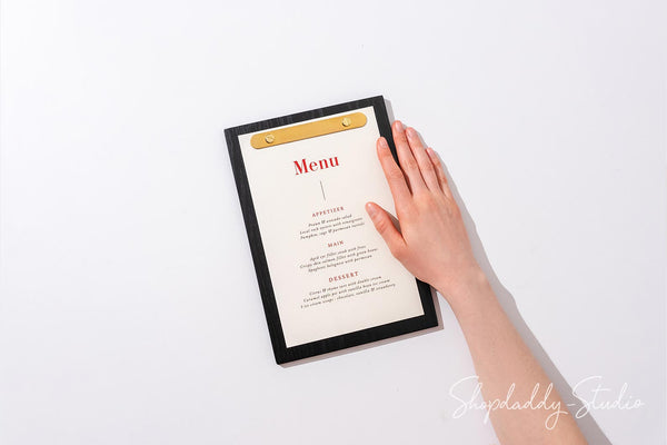 A black-framed menu board with a gold clip holding an off-white paper menu, shown from above on a white background with a hand resting on the right side. A Shopdaddy Studio watermark is visible in the bottom right corner.
