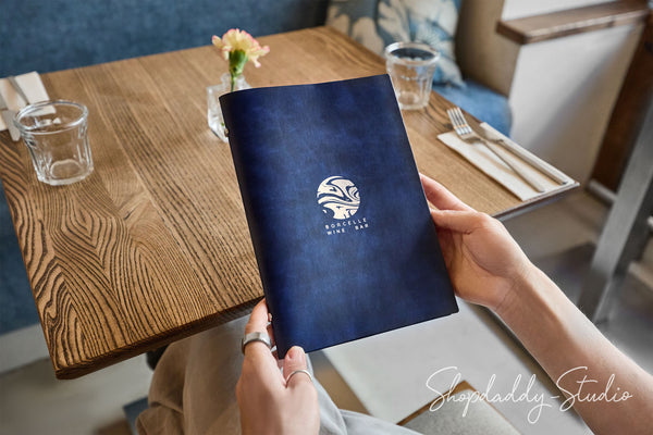 A person holding a dark navy blue menu or booklet with a white circular logo that reads 'SUNSET LAKES RESTAURANT' in a restaurant setting with a wooden table and glassware.