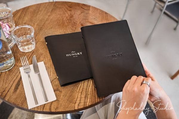 Two black leather-bound booklets with the word 'HUNGRY' printed in white on the covers are placed on a round wooden table. One is being held by hands wearing a silver ring, with a fork and knife on a napkin beside it and a glass of water nearby.