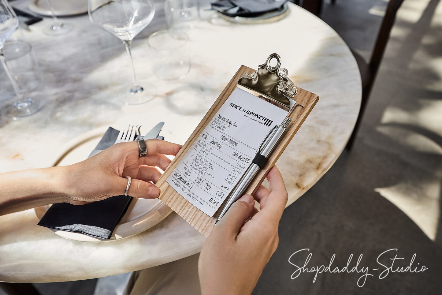 A close-up lifestyle shot of a person holding a wooden clipboard with a restaurant check print at a round marble dining table. The person's hands are visible, along with wine glasses and cutlery in the background.