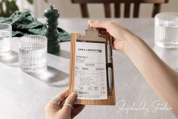 A person holds a wooden clipboard with a printed receipt and pen in a restaurant setting. The clipboard has a light wood grain finish and a silver metal clip.