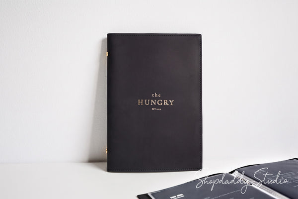 A black leather journal standing upright on a white surface against a white wall, with gold foil text reading 'the HUNGRY' and 'EST. 2014' on the cover.