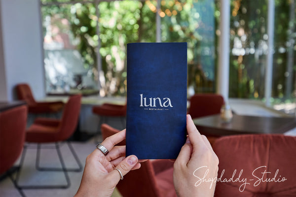 A person holds a navy blue menu or booklet with the word 'luna' printed in white in the center of the frame, with a blurred cafe interior featuring red chairs and large windows in the background.