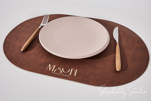 An oval brown leather placemat on a light gray background, styled with a beige ceramic plate, a wooden-handled fork on the left, and a matching knife on the right. The placemat features the brand name 'MOON' printed in gold near the bottom.