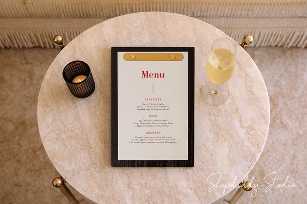 A framed vertical menu with a black frame and gold clip, displayed on a round marble table alongside a candle holder, a glass of champagne, and a decorative background.