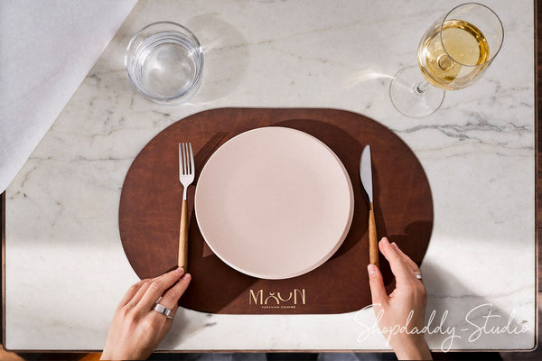 Top-down view of a place setting on a white marble table, featuring a dark brown oval placemat with the name 'MAYUN' printed in gold at the bottom, a white ceramic plate, a fork with a gold handle, a knife with a gold handle, and two hands holding the cutlery. A glass of white wine and a glass of water are also visible.