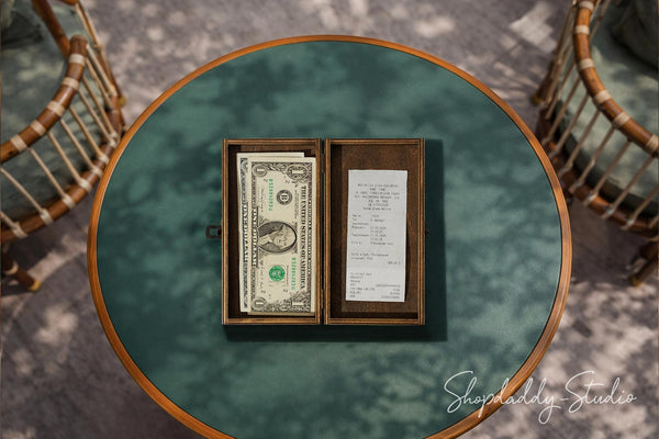 An overhead shot of a round teal-green table with a wooden rim, featuring an open dark wood box containing a stack of one hundred dollar bills and a handwritten receipt. Two woven rattan chairs are partially visible in the background on a gray patterned floor.