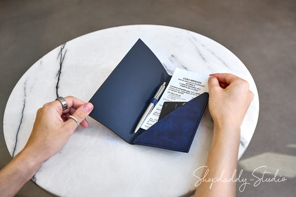 A navy blue leather passport holder or document pouch is shown open on a round white marble table, with a pen and papers inside. Two hands are visible holding the item, and a Shopdaddy Studio watermark appears in the lower right corner.