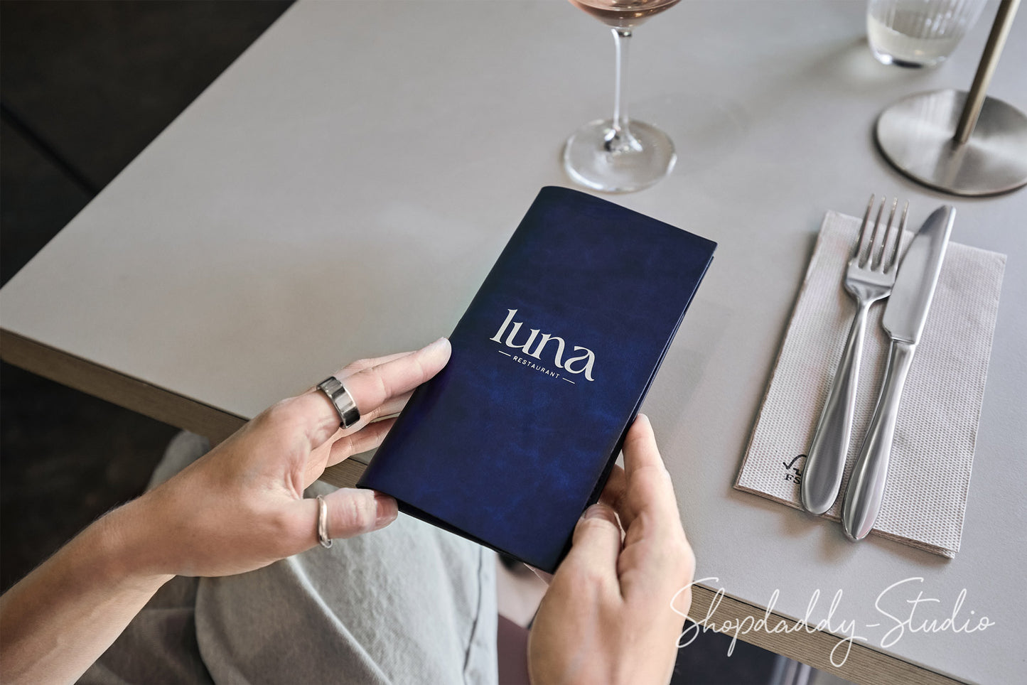A person holds a dark navy blue menu or booklet with the word 'luna' printed in white at a restaurant table setting, with a wine glass, fork, knife, and napkin visible on the table.