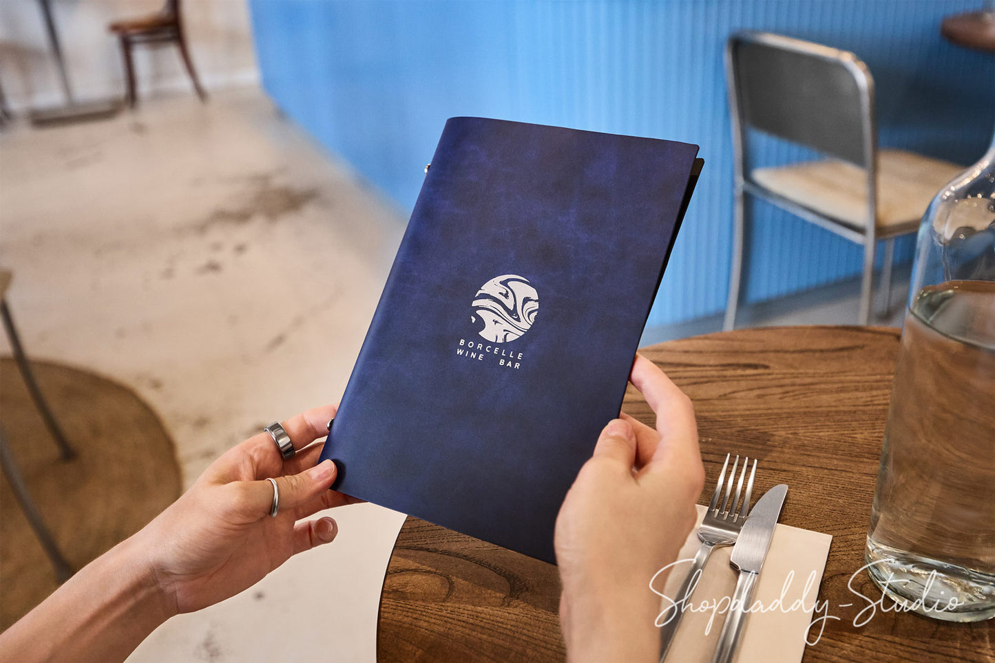 A person holds a dark navy blue menu with a white printed logo and text in a restaurant setting. The scene includes a wooden table, silverware, a glass bottle, and chairs in the background.