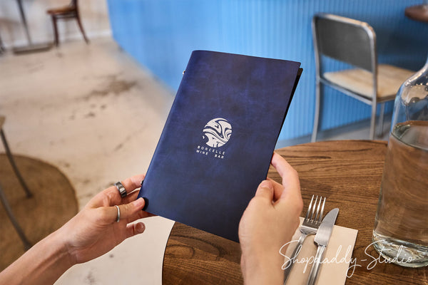 A person holds a dark navy blue menu with a white printed logo and text in a restaurant setting. The scene includes a wooden table, silverware, a glass bottle, and chairs in the background.