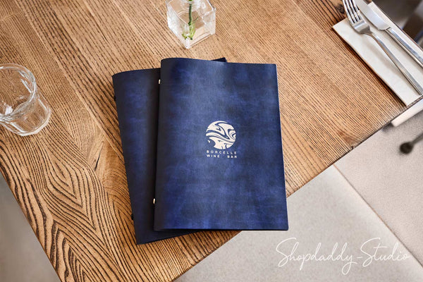A dark blue textured leather-bound menu folder with a white circular logo reading 'BUTTERFLY WINE BAR' lies on a wooden table, surrounded by dining props like a glass, silverware, and a napkin.