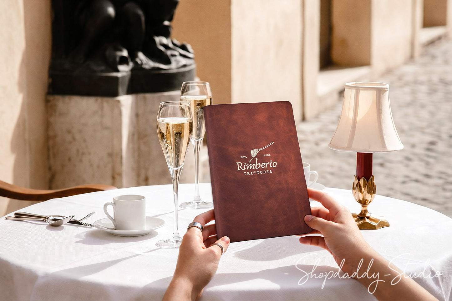 A person holds a brown leather-bound restaurant menu labeled 'Rimberto Trattoria' at an outdoor table setting with two champagne flutes, a white coffee cup, a table lamp, and warm sunlight.