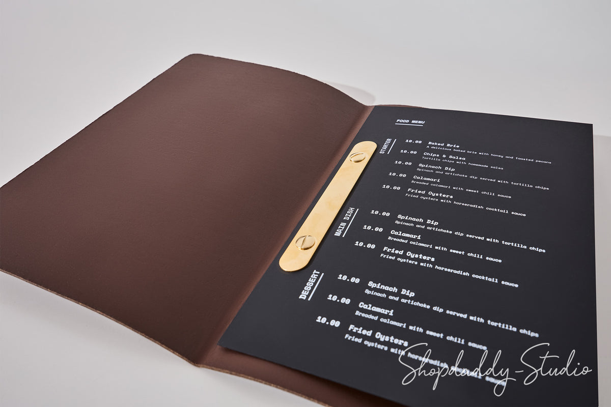 An open menu folder with a dark brown leather cover and a black printed inner page, photographed on a light gray background. The page contains white text and a gold metal clip holding the insert.