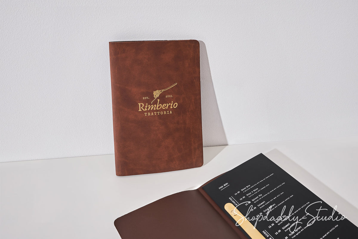 A brown leather-bound notebook standing upright, with its cover printed in gold text reading 'Rimberio' and a small bird logo. An open matching notebook lies in the foreground, showing a dark interior page with white text and a gold bookmark.
