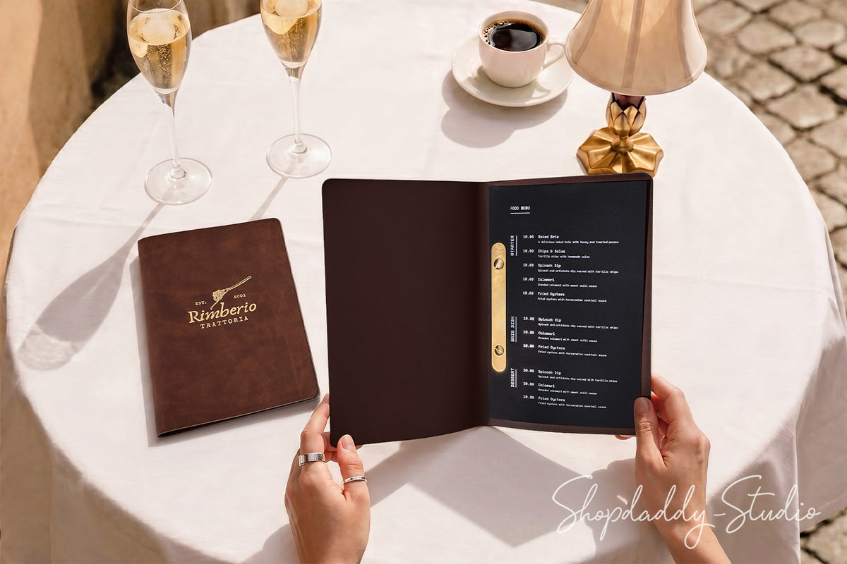 A person holds an open dark brown leather-bound restaurant menu with a gold ring binder, set on a white tablecloth beside a closed matching menu, two champagne flutes, a coffee cup, and a lamp.