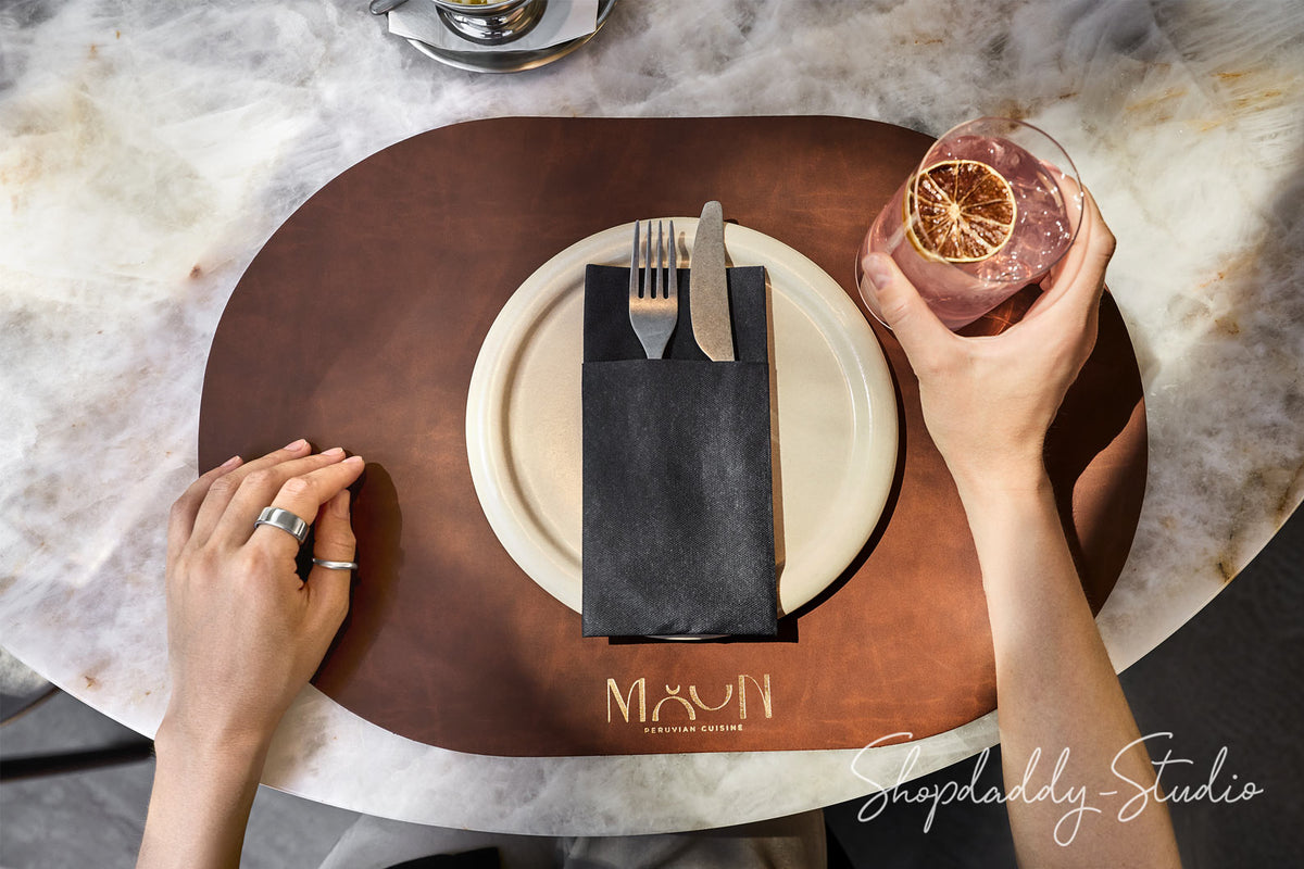 Overhead view of a dining table setting featuring a brown leather placemat with the gold logo 'MAON EST. 2018', a white plate, a black napkin holding silver cutlery, and a hand holding a pink cocktail with a dried orange slice.