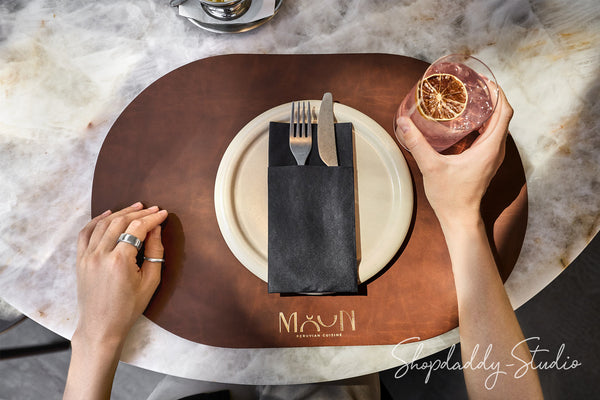 Overhead view of a dining table setting featuring a brown leather placemat with the gold logo 'MAON EST. 2018', a white plate, a black napkin holding silver cutlery, and a hand holding a pink cocktail with a dried orange slice.