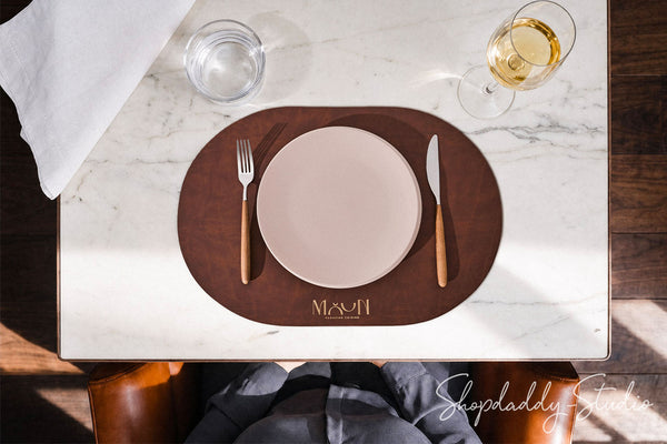 An overhead view of a styled dining table setting featuring a dark brown oval placemat with the name 'MAXHN' printed in gold at the bottom, accompanied by a beige plate, wooden-handled fork and knife, a glass of white wine, and a glass of water on a white marble tabletop.