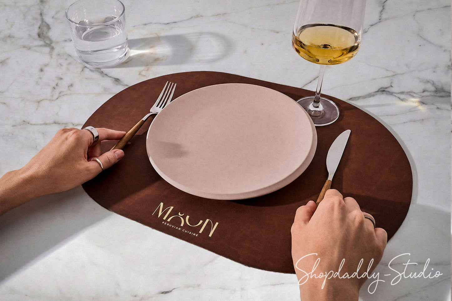 A brown oval placemat on a white marble table, featuring the gold logo 'MOON luxury dining'. A pale pink ceramic plate is placed on top, with a fork on the left and a knife on the right, held by two hands.