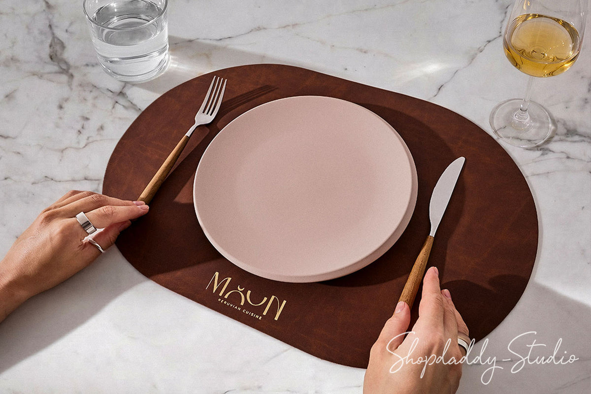A styled dining setup on a white marble table featuring a dark brown oval placemat with gold lettering, a pale pink plate, a fork with a wooden handle, a knife with a wooden handle, and hands holding the utensils. A glass of water and a glass of white wine are also visible.