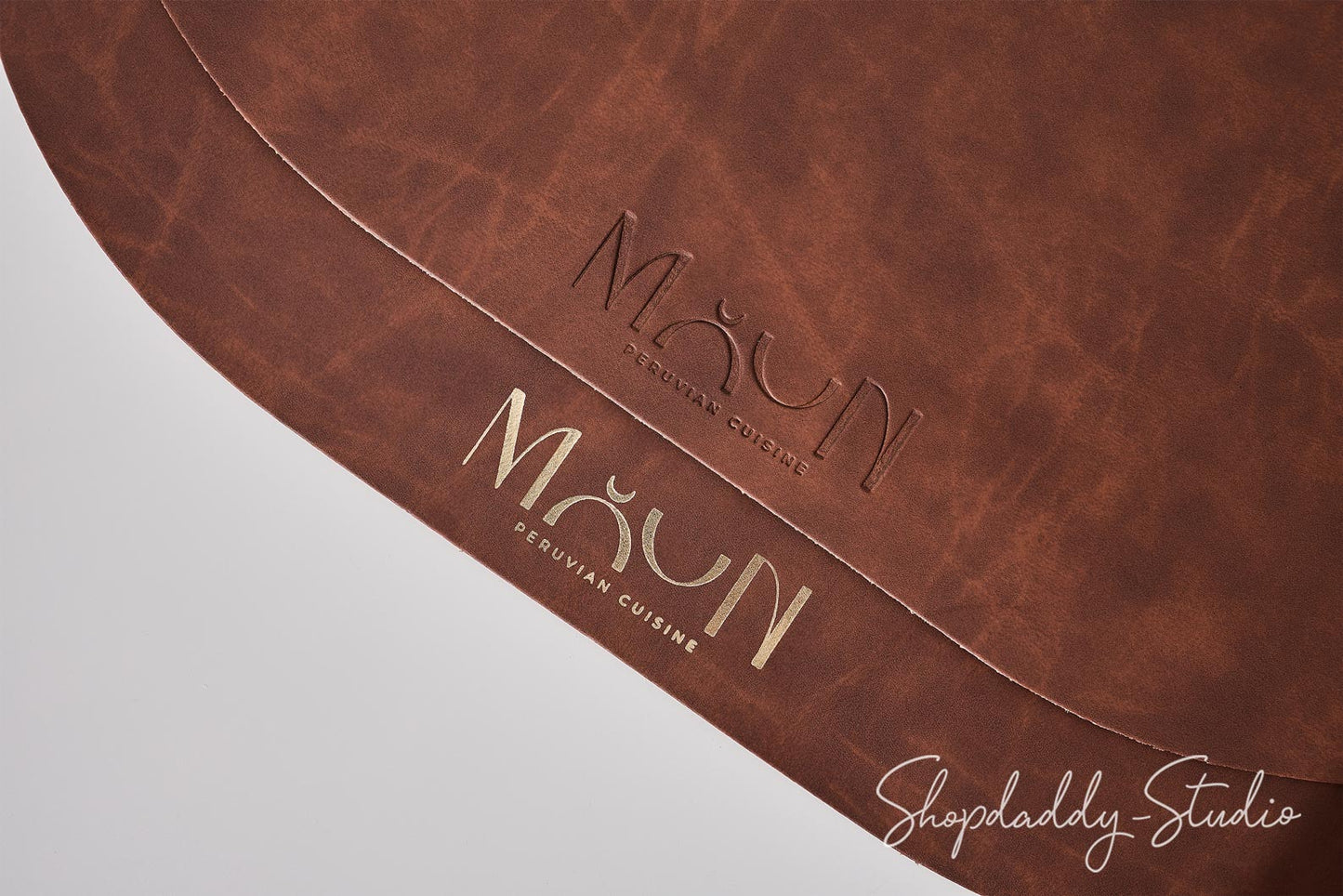 A close-up of a brown leather or faux-leather surface with the brand name 'MAUN PERuvian Cuisine' embossed in the center and gold-toned lettering near the bottom. A Shopdaddy-Studio watermark appears in the lower right corner.