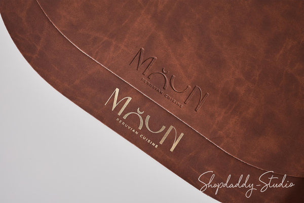 A close-up of a brown leather or faux-leather surface with the brand name 'MAUN PERuvian Cuisine' embossed in the center and gold-toned lettering near the bottom. A Shopdaddy-Studio watermark appears in the lower right corner.
