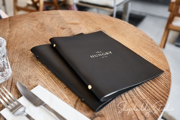 Two black leather-bound menus with gold lettering reading 'the HUNGRY' stacked on a round wooden table, with a glass, fork, and knife nearby.