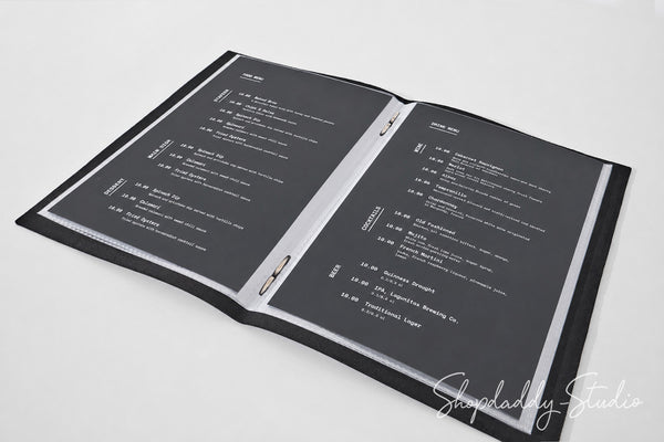 An open black binder or menu folder with two dark gray pages printed with white text, photographed on a light gray background. A script watermark reading Shopdaddy-Studio appears in the lower right corner.