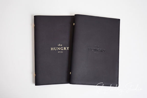 Two black leather book covers or portfolios with gold lettering reading 'the HUNGRY by JEN' are laid flat on a white background.