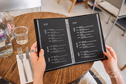 A person holding an open black printed menu at a round wooden table in a cafe setting, with hands, cutlery, glasses, and chairs visible in the background.
