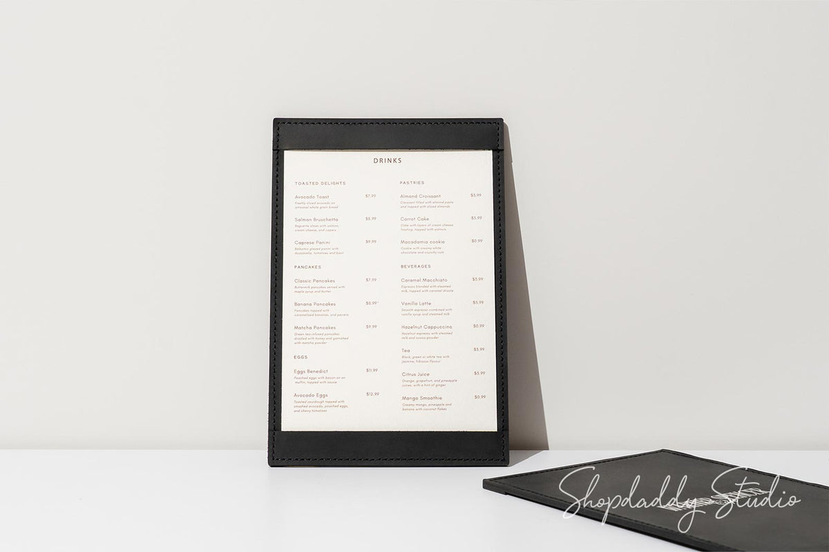 A black leather menu holder standing upright on a white surface against a white wall, displaying a printed menu insert. A second black menu holder lies closed on the table in the foreground.
